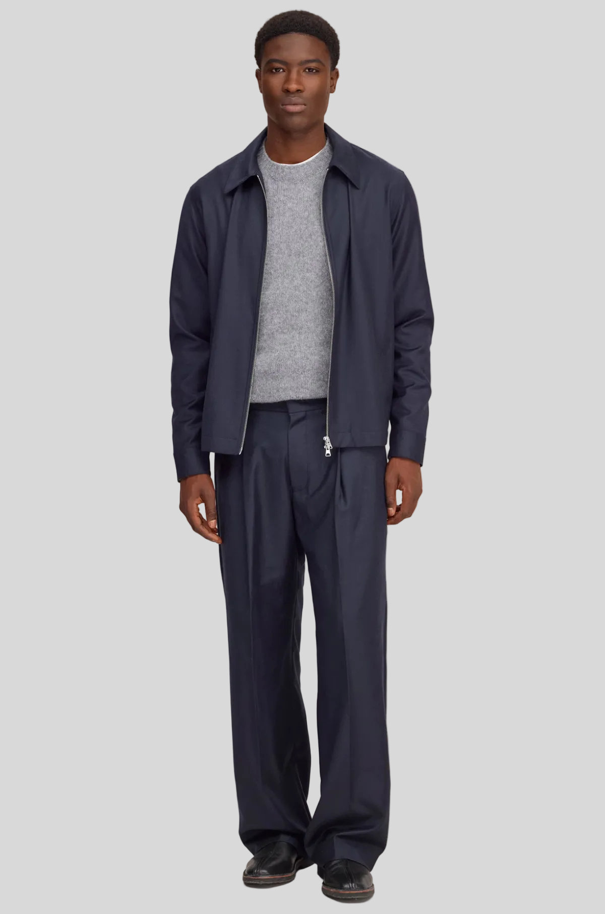 A man modeling a navy blue lightweight jacket and matching trousers over a gray knit sweater, standing against a solid light beige background.