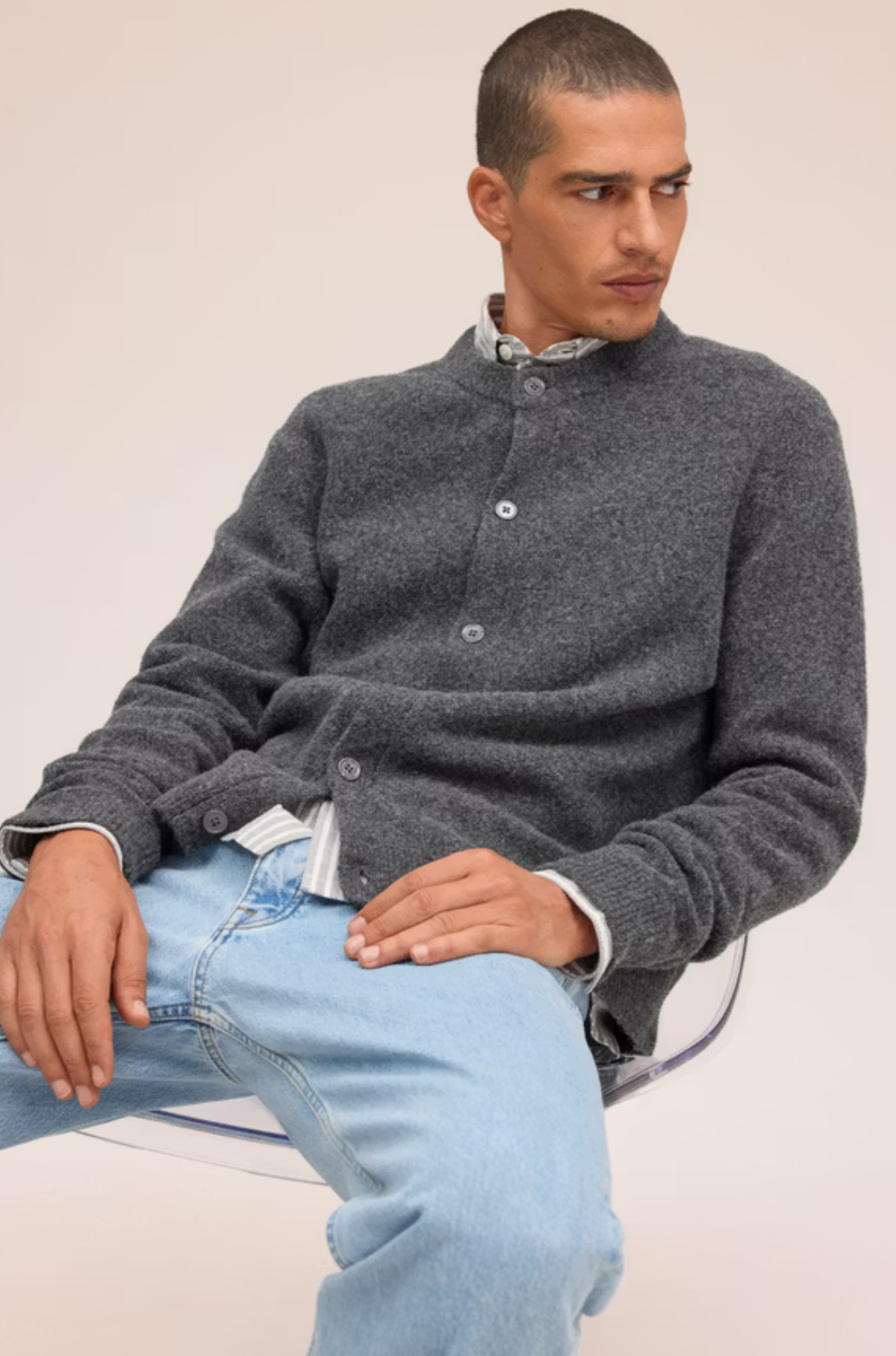 A man wearing a dark gray heathered knit cardigan over a light blue and white patterned collared shirt, paired with light wash blue jeans, seated against a plain beige background.